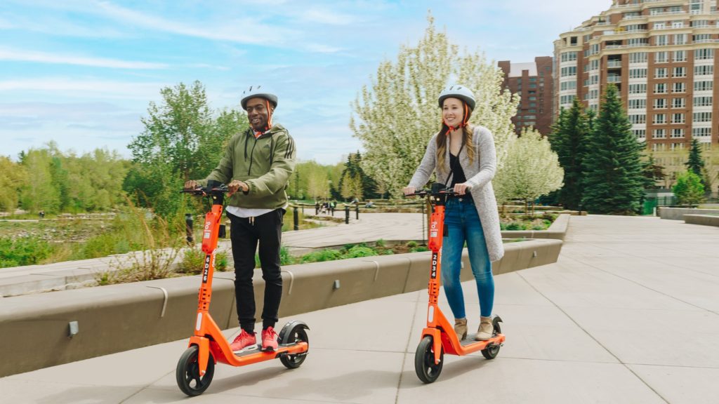 People riding Neuron bullfrogpowered scooters in a green city
