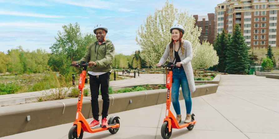 People riding Neuron bullfrogpowered scooters in a green city